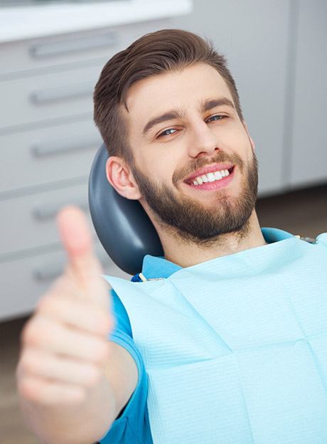 Young bearded man in dental chair wearing bib smiling and giving thumbs-up to oral care. Dental cleanings, dental hygiene, oral health, white teeth.